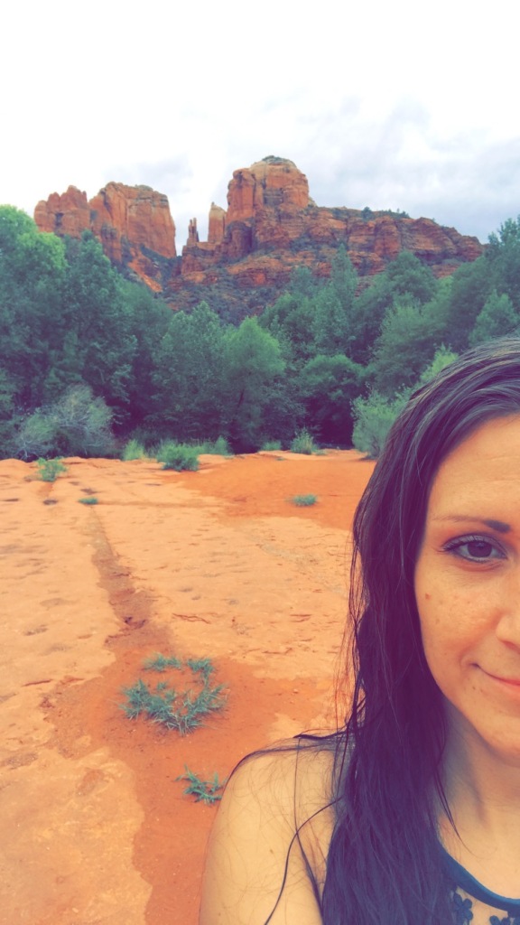 A smiling woman standing in front of the red rocks of Sedona with green trees outlining the base of the mountain.